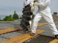 Osoby w kombinezonach ochronnych usuwają eternit z dachu. Koszt usunięcia azbestu zależy od wielu czynników.