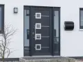 Nowoczesne drzwi zewnętrzne w ciemnoszarym kolorze z geometrycznymi przeszkleniami i metalowymi elementami. Obok skrzynka na listy.
