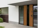 Nowoczesne, drewniane drzwi wejściowe z czarnym panelem i przeszkleniem. Obok okno z żaluzjami i zielona roślinność.