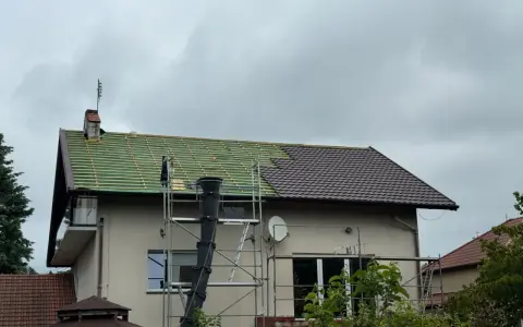 Remont dachu domu: połowa pokryta zieloną łacą, połowa brązową dachówką, rusztowanie.