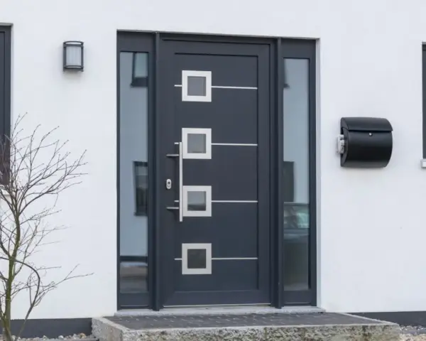 Nowoczesne drzwi zewnętrzne w ciemnoszarym kolorze z geometrycznymi przeszkleniami i metalowymi elementami. Obok skrzynka na listy.