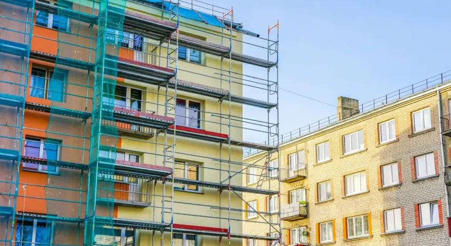 Budynek w remoncie, rusztowania i siatka ochronna. Czy balkon jest częścią mieszkania? Widać różne balkony, niektóre z czerwonymi markizami.