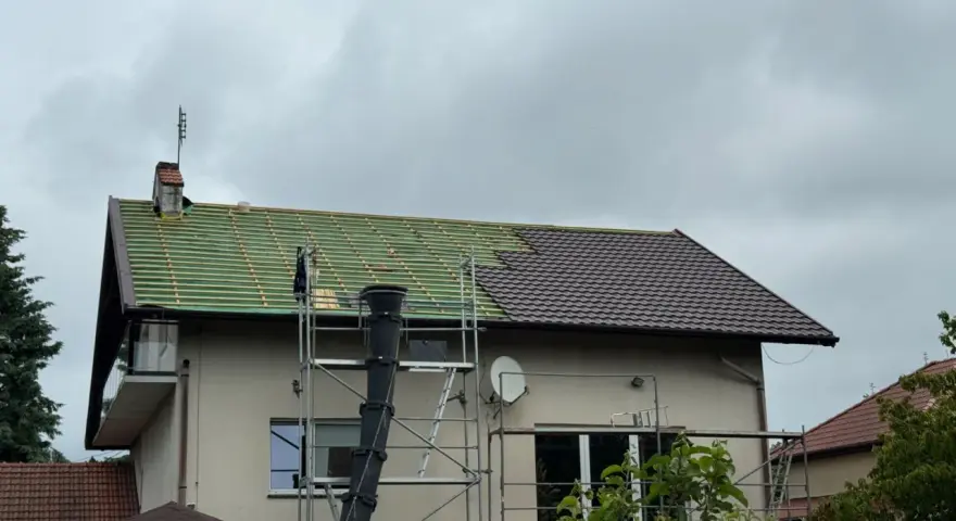 Remont dachu domu: połowa pokryta zieloną łacą, połowa brązową dachówką, rusztowanie.