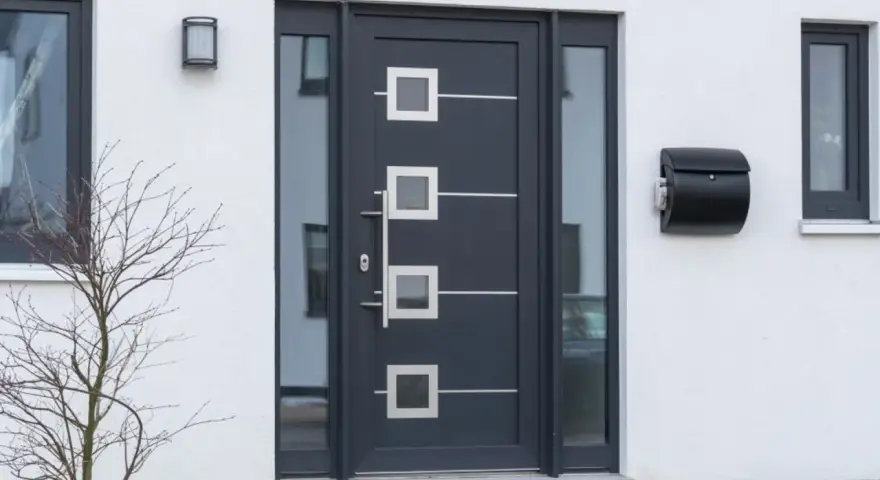 Nowoczesne drzwi zewnętrzne w ciemnoszarym kolorze z geometrycznymi przeszkleniami i metalowymi elementami. Obok skrzynka na listy.