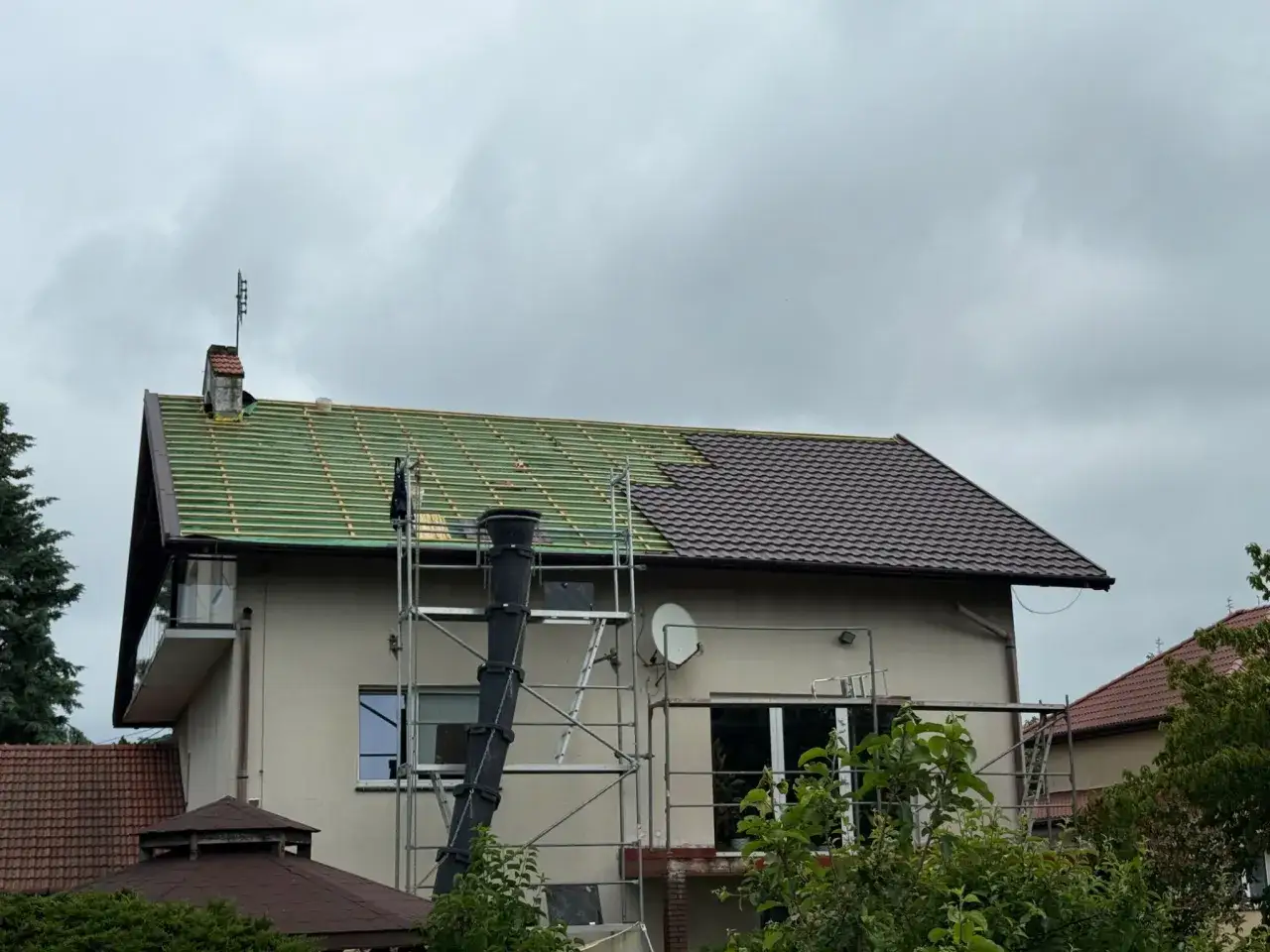 Remont dachu domu: połowa pokryta zieloną łacą, połowa brązową dachówką, rusztowanie.