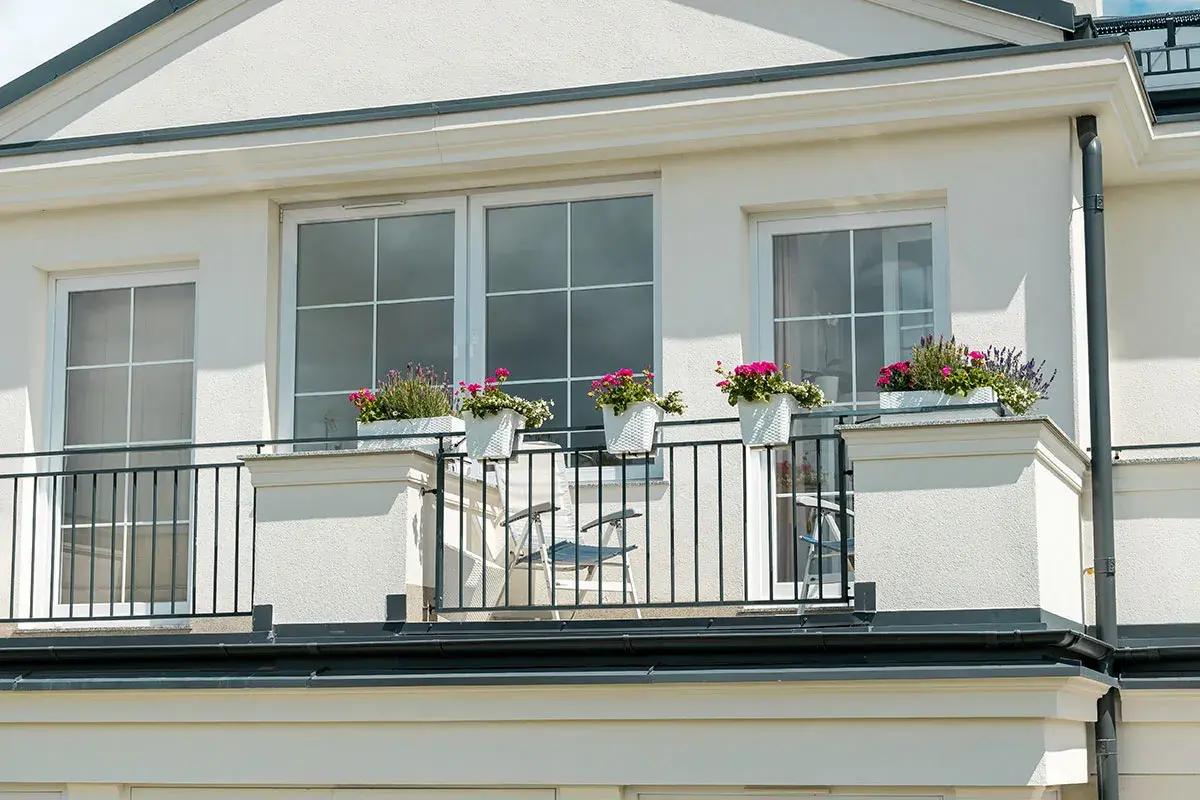 Białe okna i jasna elewacja domu z brązowym dachem. Balkon ozdobiony donicami z kwiatami.